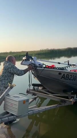 A man in camouflage clothing is pushing a boat that is on a trailer into a body of water. The boat is loaded with gear, including what appears to be fishing equipment and life vests. The trailer is partially submerged in the water, and the boat is being guided off it. The water is calm and reflects the clear sky and the surrounding vegetation. The scene appears to be an outdoor recreational setting, likely a lake or river, with reeds lining the bank in the background.