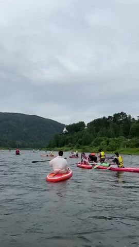 The video shows a group of people on stand-up paddleboards on a river. The sky is cloudy and overcast. There are trees and a hill in the background. The people are spread out on the water, some paddling and some sitting on their boards. The person in the foreground is on a red paddleboard, facing away from the camera. Other paddleboards are visible in the background, some with multiple people. The water is rippling, indicating a gentle current. There is a small white building visible on the hill in the background. The overall impression is of a recreational outing on a river.