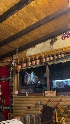 The video shows a room with a rustic aesthetic. A row of copper-colored bells hangs from a wooden beam, secured with brown leather straps. Above the bells, a piece of white sheepskin or a similar textured fabric is mounted on the wall. The ceiling is made of wooden planks. Below the bells, there's a window reflecting a television screen showing a fireplace. A sign with writing in a foreign language is visible, along with other decorative items such as flower arrangements and ornaments. The room has a cozy and folksy ambiance. A deer skull with antlers is hung to the left. The video moves closer to the copper bells.