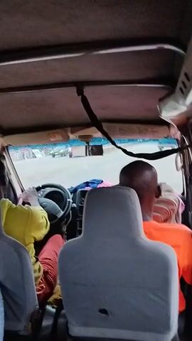 The video shows the interior of a vehicle, possibly a van, from a rear passenger's perspective. The camera is pointed forward, looking towards the driver and the windshield. Several people are visible inside the vehicle. In the driver's seat, a person wearing a yellow shirt and red pants is seen, with their hands on the steering wheel. To the right of the driver, another person with a shaved head and wearing an orange shirt is visible. In the foreground, the backs of two headrests of passenger seats are prominent. Through the windshield, other vehicles and a road are visible, suggesting the vehicle is in motion. The overall lighting and visual quality indicate the video was likely taken during daytime.