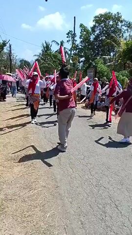 A group of people are parading down a paved road, holding red and white poles. They are wearing white shirts, red headscarves, and black pants. Some are also wearing checkered skirts. The road is lined with trees and buildings. The sky is blue and there are shadows on the ground. There is a green umbrella in the top left corner of the frame. A person in a maroon shirt and tan pants is walking away from the camera, facing the parade.
