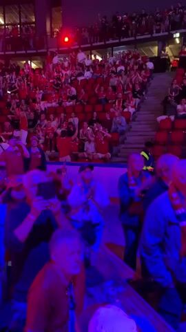 The video shows a crowd of people in an arena. The people are mostly facing forward, towards a stage or playing area that is not fully visible. Many are wearing red shirts and scarves, suggesting they are fans of a sports team or musical group. Some are waving their arms and appear to be cheering or singing along. The lighting is dim, with blue and pink lights illuminating the crowd. There is a large screen visible in the background, displaying a colorful and dynamic image. The environment appears to be a large indoor venue, possibly a stadium or concert hall.