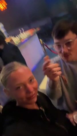 Two people are at a bar, taking a selfie. The woman in the foreground has short blonde hair and is wearing a black jacket. She is smiling widely, holding a dark beer in her right hand. Behind her, a man with brown hair and glasses makes exaggerated gestures with his hands, pointing at the camera and making faces. He wears a light grey sweatshirt. The bar in the background has a white counter with colorful lights and bottles. The overall lighting is dark with neon accents. The camera is shaky, suggesting it's being held by one of the people in the shot. Music plays in the background.