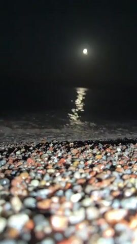 The video shows a low-angle view of a pebble beach at night, with the moon visible in the dark sky. The moon is casting a reflection on the dark water, creating a shimmering path. The waves are gently lapping at the shore, covering the pebbles. The focus is on the pebbles in the foreground, which are wet and glistening. The overall atmosphere is dark and serene.