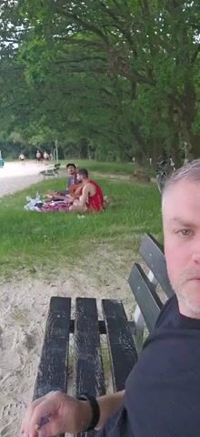 The video shows a man taking a selfie in the foreground, sitting on a wooden bench. Behind him, on a sandy area and grassy bank, a group of people are relaxing. Two men are sitting on a blanket, one playing a guitar, and another man is visible to their left. In the background, several other people are walking near the water. The setting appears to be a park or beach area with trees and foliage surrounding the scene.