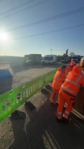 The video shows a group of construction workers in bright orange work suits at a construction site on a sunny day. Three workers are standing around a large, round wooden spool, talking, and appear to be on a break. The ground is gravel, and there are several other spools of various sizes nearby. A green and white plastic fence surrounds the work area, and in the background, there is a large parking lot filled with various vans, trucks, and other construction vehicles. Beyond the vehicles, there is a metal fence and some trees. High-tension power lines stretch across the sky, and a large electrical tower is visible. A worker in an orange suit walks past the spool towards the right side of the frame.
