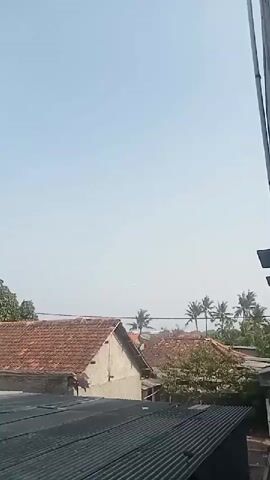 The video shows a wide view of a residential area under a clear blue sky. In the foreground, a corrugated metal roof is visible. Behind it, several houses with terracotta tile roofs are shown. Trees, including some palm trees, are scattered throughout the scene. The overall impression is one of a sunny day in a somewhat rural or suburban setting. There are no people or discernible actions taking place in the video.