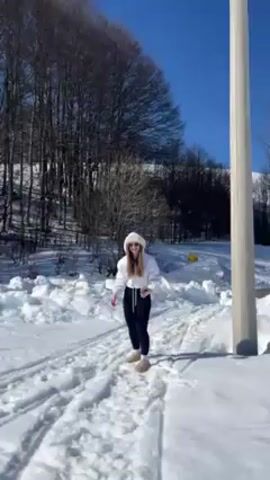 The video shows a young woman in a snowy environment. She is wearing a white hooded sweatshirt, black leggings, and light-colored slippers. The woman is standing on a snow-covered path with tire tracks. Behind her is a snowy hillside with bare trees, and the sky is blue.

The woman begins by tossing something up in the air, then it appears she drops her cell phone in the snow. She bends down to pick it up.