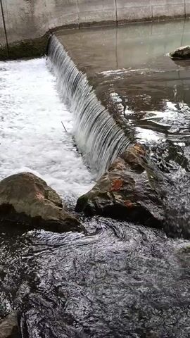 The video shows a close-up, slightly angled shot of a small waterfall or dam. Water cascades over the edge, creating white foam and spray. Rocks are visible on both sides of the falling water, with the river below appearing dark and rippled. The background shows a concrete structure, possibly the edge of a canal or man-made waterway. The lighting suggests it's daytime, possibly overcast, with reflections on the water surface. The focus is on the movement and texture of the water and rocks.