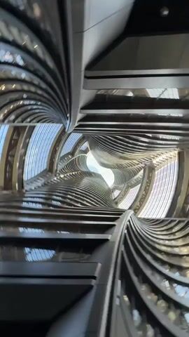 The video presents a low-angle, dynamic view looking directly upwards through a complex architectural structure, likely the interior of a tall building. The structure features a series of curving, horizontal planes or balconies stacked vertically, creating a vortex-like or twisting effect as the eye travels upward. These planes are metallic or gray in color, and their edges create a rhythmic pattern of lines that converge toward a central opening at the top, revealing the sky. The building is likely contemporary in design, emphasizing geometric patterns and open spaces. The camera movement is fluid, adding to the disorienting and visually engaging perspective. The overall environment is modern and architecturally intricate.