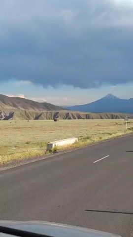 The video shows a view from a vehicle moving along a paved road. The road stretches ahead, with white lines marking the lanes. To the left of the road, there is a dry, grassy field with some hills in the distance. A small, dark brown structure is visible in the field. Further in the distance, a large, snow-capped mountain dominates the horizon, with another smaller mountain to its left. The sky is partly cloudy, with dark storm clouds gathering in the upper left portion of the frame. The overall environment appears to be a rural, mountainous landscape.