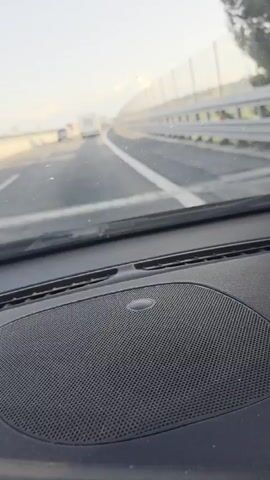 The video shows a first-person view from inside a vehicle traveling on a highway. The camera is positioned looking forward through the windshield. The road ahead is paved with a white line marking the lane. To the right, there is a guardrail and a fence that extends into the distance, separating the highway from a rural landscape. Several vehicles are visible on the road, including trucks and cars. The sky is clear and bright, indicating daytime. The interior of the vehicle, including the dashboard and a speaker grille, is visible in the foreground. The vehicle appears to be in motion, as indicated by the slight blur and the changing perspective of the road and surrounding environment.