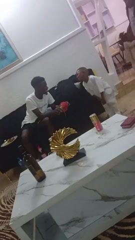 In a brightly lit room, a young man in a white t-shirt and shorts sits on a black sofa and plays with a red and green object, while a small boy in a white outfit stands nearby, leaning on the sofa. Another young girl, wearing a dress, is visible in the background. A white marble coffee table with a golden sculpture, a can, a wallet, and a bottle rests in front of the sofa. The rug beneath the coffee table is patterned. A painting hangs on the wall, and a partially visible dining area with chairs is in the background. The boy playfully hits the sofa with his hand.