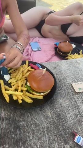 Two individuals are seated at a wooden table on a sandy beach. In the foreground, a black plate contains a hamburger and french fries. To the left, another person is reaching for their plate, which also has a hamburger and french fries. The person on the right is wearing a black bikini and sunglasses, with a burger and fries on a plate in front of them. A pink towel and a cell phone are also visible. The environment appears to be a beach with wooden walkways and beach chairs in the background.