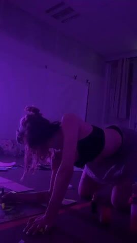 A young woman with her hair in a bun is on her hands and knees on a floor that appears to be a studio or practice space. She is wearing a black sports bra and light-colored shorts. The room is lit with purple light. She is holding what appears to be a marker and drawing on a large piece of paper spread out on the floor. She then stands up and jumps into the air, arms outstretched. She lands on her knees. In the background, there are curtains, a whiteboard, and some objects on the floor, including bottles and tape.