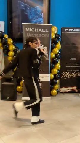 The video shows a male performer dressed as Michael Jackson, wearing a black suit with white accents and white fur cuffs on his socks. He is standing in front of a banner that says "MICHAEL JACKSON" with a picture of the singer. To his right, there is another banner with a poster and the date "23 DE ABRIL". The performer is holding a microphone and appears to be singing or talking. He is also performing some dance moves, including a side-step and a kick. There are black and gold balloons on either side of the banner. In the background, there is a poster for "MX4D" and a blue wall. Another person, wearing a black t-shirt and jeans, stands to the right of the performer, watching him. The floor is tiled.