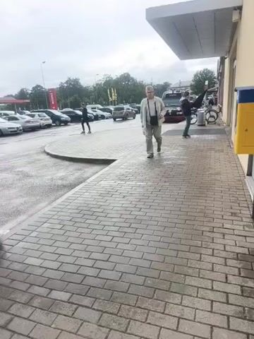 A man is walking towards the camera on a brick walkway. He is wearing glasses, a light-colored jacket, and light-colored pants. He is carrying a bag over his shoulder and another bag in his hand. Behind him, a parking lot is visible with several cars. A red truck and a bicycle are also in the background. The sky is overcast.