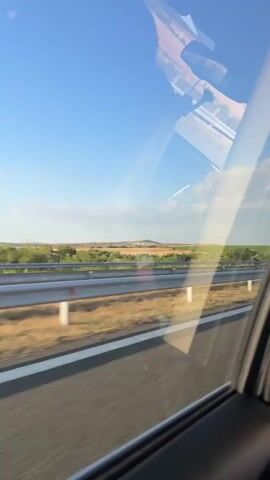 A first-person perspective video shows a view from inside a vehicle, looking out the passenger window. The vehicle is traveling on a highway with a guardrail visible in the foreground. The road has white lane markings. Beyond the guardrail, there are fields of dry grass and green crops. In the distance, a small hill with what appears to be communication towers on top is visible under a clear blue sky with scattered clouds. The reflection of the vehicle's interior, including a dashboard and possibly a passenger, is visible on the car window. The motion blur indicates the vehicle is in motion.