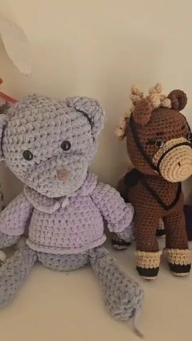 A collection of handmade crocheted stuffed animals is displayed on a shelf. On the left, a large grey teddy bear wearing a lavender hoodie is positioned. To its right, a small brown horse with a tan mane and halter stands. Next to the horse is a purple elephant with pink ears and feet. Further to the right, a white bunny wearing a maroon hat and dress with a yellow skirt is seated. Several crocheted objects, including a brown egg-shaped item, are also visible. The background is a plain, light-colored wall.