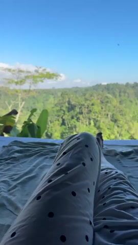 The video opens with a close-up of a person's legs, clad in white pajama pants with black polka dots, resting on a bed covered with a light-colored sheet. The perspective is from the person's point of view, looking forward. The background reveals a lush, green jungle landscape under a clear blue sky with a few white clouds. A bird is visible flying in the upper right portion of the frame. The camera slowly pans upwards, offering a wider view of the dense tropical forest stretching out to the horizon. The lighting suggests it is daytime, likely morning or afternoon.