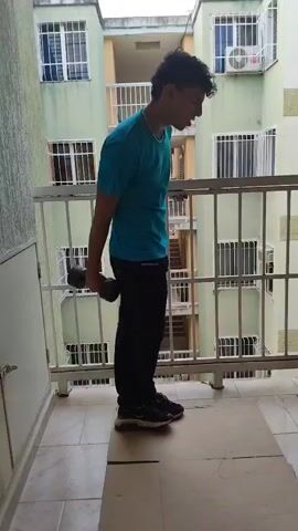 A person stands on a balcony, performing dumbbell exercises. The person is wearing a blue t-shirt and black pants. They are holding dumbbells in each hand and move them in a circular motion in front of their body. The balcony has a white railing and overlooks a building with green walls and windows.