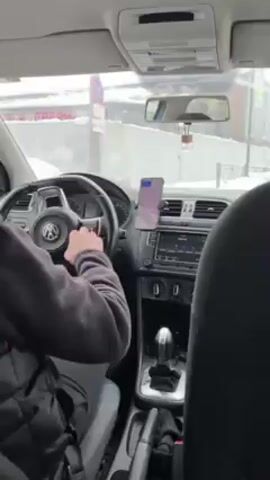 The video shows the interior of a car, seemingly a Volkswagen Polo, from the perspective of the back seat. A person wearing a black puffer vest is driving. The phone is mounted on the dashboard, and a gear stick is visible. Outside the car, snow is visible. The video briefly shows the driver's head and back. A blue bag is on the back seat. The camera is focused on the interior of the car and the driver. The car appears to be stationary.