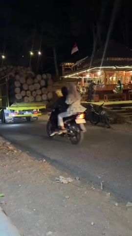 A nighttime scene unfolds on a paved road. In the foreground, the road surface is rough and appears to be under construction or in disrepair, with loose gravel and dirt visible. A curb runs along the left side of the road. In the background, a truck laden with large logs is parked, its headlights illuminated. Beside the truck, a motorcycle carries two individuals, a driver and a passenger, both wearing head coverings. The motorcycle is in motion, heading away from the camera. To the right of the road, an elevated structure resembling a restaurant or cafe is illuminated by string lights. Several motorcycles are parked near this structure. A black pickup truck with its brake lights illuminated passes by the camera, moving in the same direction as the motorcycle. The overall environment suggests a rural or semi-rural setting at night, possibly a mountain area given the presence of logs and the elevated structure.