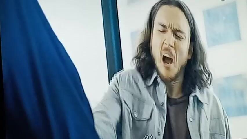 The video opens with a man with long, dark hair and a light-colored button-down shirt grimacing. The scene then shifts to a man wearing a blue hoodie with the hood up, looking to the side with a beard. He is wearing a light-colored shirt underneath. In the next shot, a close-up shows a person's hands holding a blue smartphone connected to a charger, with a keyboard and mouse visible in the background. The phone screen then displays a video call with a woman who has bright pink hair and is smiling.