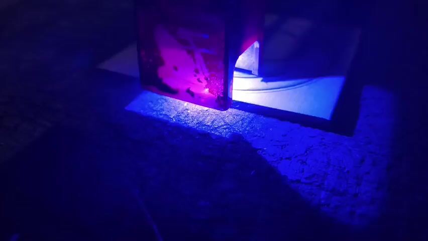 The video shows a close-up, low-light shot of a red, translucent cube placed on a wooden surface. A thin, white piece of paper or card is positioned beneath the cube. A bright blue light emanates from beneath the cube, illuminating the area. The cube itself glows with a purple hue, and within it, indistinct shapes or contents are visible. The blue light casts shadows on the wooden surface and the paper. The camera appears to be mostly stationary, with slight movements or vibrations. The overall impression is of a scientific or experimental setup, possibly involving fluorescence or light refraction. There are no people visible in the video.