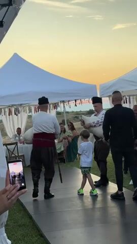 A vertical outdoor shot captures a vibrant evening event. Two men in traditional clothing, one playing a drum and the other a bagpipe, stand on a black runway. A young boy in a white t-shirt and camouflage shorts stands between them, watching intently. To their left, several women in dresses are seated at a table under a white canopy, observing the performance. Another man in a black shirt stands to the right of the performers. The scene is set against a backdrop of a clear sky with an orange sunset. In the foreground, the lower part of a person holding a phone is visible, capturing the moment.