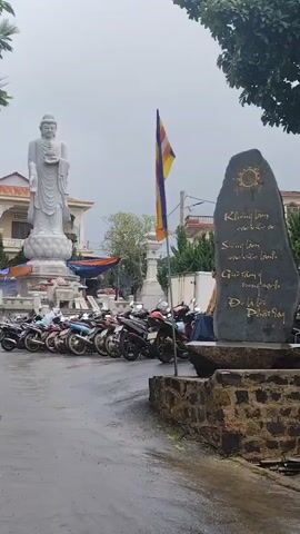 A standing Buddha statue is visible in the background, with a Buddhist flag flying nearby. A row of motorcycles is parked in front of the statue, and a man in a tan outfit is walking past them. The ground is wet, suggesting recent rain. A large stone monument with inscriptions and a sun symbol is in the foreground. The overall atmosphere is subdued, possibly due to the overcast weather.