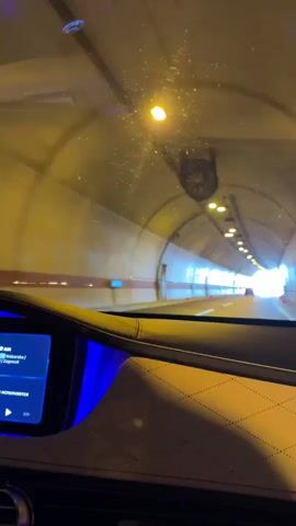 The video shows a first-person view from inside a car driving through a tunnel. The tunnel is lit by overhead lights and has concrete walls. The car's dashboard is visible in the foreground, with a screen displaying what appears to be music information. The road ahead is visible, with a white line marking the lanes. The tunnel exit is visible in the distance as a bright light. The overall impression is of a modern highway tunnel.