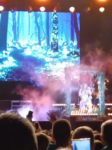 The video shows a singer performing on stage at a concert. She is sitting on a swing decorated with greenery, and a large screen behind her displays an image of a magical forest with glowing plants. The stage is lit with bright spotlights and filled with fog. The audience, visible in the foreground, is holding up their phones to record the performance. The singer is dressed in a light-colored outfit, likely a dress, and is holding a microphone.