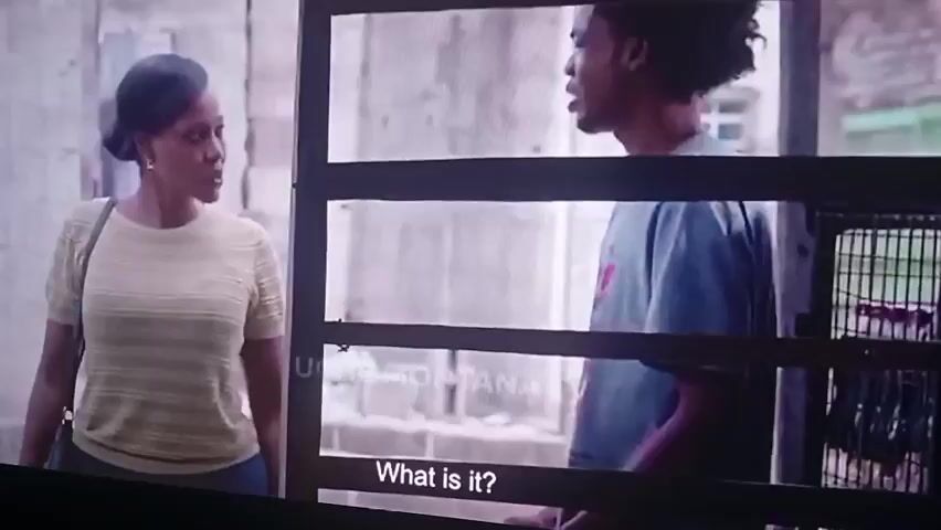 The video shows a scene with two people. On the left, a woman with dark hair pulled back is wearing a light-colored, textured top and dark pants. She is looking towards the right. On the right, a young Black man with curly hair is wearing a gray t-shirt with a pink graphic. He is looking towards the left. The background appears to be an outdoor urban setting with some fencing and buildings. Subtitles are visible at the bottom of the screen. The first subtitle reads "What is it?". The second subtitle reads "It's Daddy".