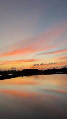The video shows a body of water reflecting a sunset sky with scattered clouds. The sky is colored with hues of pink, orange, and blue. The water is still, creating a clear reflection of the sky and the silhouette of trees and land along the horizon. The viewpoint appears to be from a slightly elevated position looking across the water. There are no people or animals visible in the video.