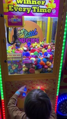 A young child plays a crane game in a brightly lit arcade. The game, “My Little Ducks,” is filled with colorful rubber ducks of varying shapes and sizes. The child, wearing a brown shirt and blue wristband, uses both hands to manipulate the controls, attempting to grab a duck with the crane claw. The machine promises a win “every time.” LED lights decorate the sides of the game.