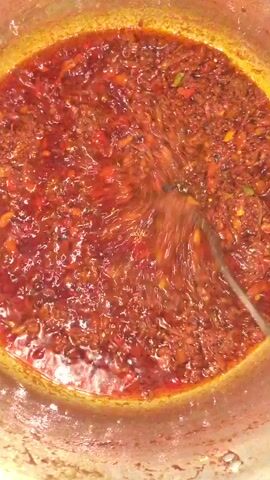 A top-down shot shows a silver metal pot filled with a dark, reddish-brown oily liquid. Small pieces of solids, including what appear to be chopped red and yellow peppers, are suspended throughout the liquid. A metal spoon stirs the mixture in a circular motion. The edges of the pot have a slight golden sheen.