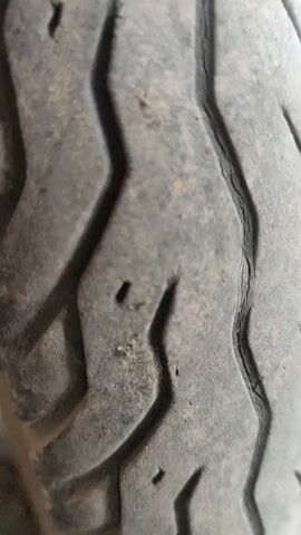 The video shows a close-up of a dirty motorcycle tire and wheel. The tire tread is visible, showing some wear and tear, including small cracks and embedded debris. The wheel is also covered in dust and dirt. A chain and other mechanical components are visible behind the wheel, also covered in grime. The tire sits on a tiled surface. The lighting is natural, possibly indoors near a window.