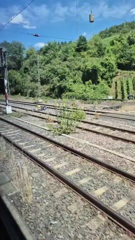 The video shows a train track with multiple sets of rails stretching into the distance. Lush green trees and vegetation cover a hillside to the right of the tracks. The sky is blue with some scattered clouds. A railway signal is visible on the left side of the frame, and several yellow posts are positioned along the tracks. The camera appears to be moving, possibly from inside a moving train, as indicated by the slight blur and the perspective. There are no people or animals visible in the video.