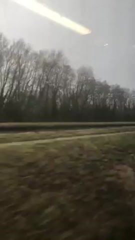 The video shows a view from a moving train, looking out the window at a blurred landscape. The landscape consists of a grassy field, a dark band that may be a road or another field, and a line of bare trees against a gray, overcast sky. The interior of the train is visible as well, including a white plastic chair.  The overall impression is one of speed and dull, gray weather.