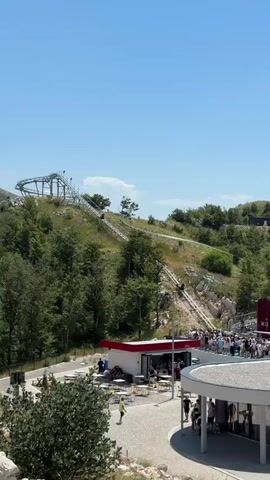 This media file depicts a bright, sunny day at an outdoor amusement attraction situated on a lush green hillside. The primary focus is a mountain coaster, a type of roller coaster where riders control their speed down a track. The track itself is visible snaking its way up and down the steep terrain, with several carts in motion.

In the foreground, there is a paved area with a modern, white building with a circular element, and a smaller red-roofed structure that appears to be a cafe or snack bar. Numerous people are gathered around these buildings and along pathways, suggesting a lively atmosphere. Some are seated at outdoor tables, while others are standing in queues or strolling. One person in a bright yellow shirt is walking across the paved area.

The background is dominated by the verdant slopes of the mountain, covered in dense trees and some rocky outcrops. The sky is a clear, vibrant blue, indicating pleasant weather. The overall impression is one of a popular tourist destination, likely a recreational park or adventure spot, offering an exciting ride experience amidst natural beauty. The time of day is clearly daytime, likely mid-morning to afternoon, due to the bright sunlight and long shadows. There is no discernible text visible in the media file.