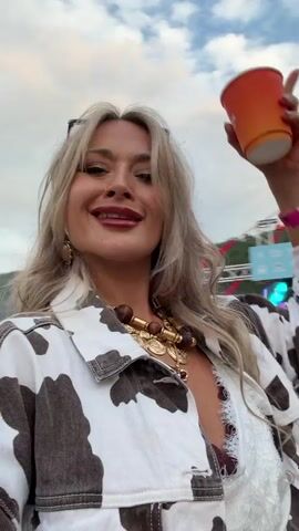 A woman with blonde hair and red lipstick, wearing a cow-print jacket and a large gold necklace, is smiling and sticking her tongue out at the camera. She is holding an orange cup in her right hand. The background appears to be an outdoor event or festival, with a stage and other people visible. The sky is cloudy.