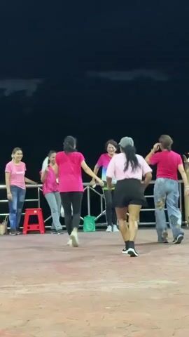 A group of six women are dancing outdoors at night. They are all wearing pink tops. The ground is paved with red and brown tiles. There is a railing behind them, and beyond that, a dark sky with some clouds. One woman in the center is wearing a pink shirt and black pants and is facing away from the camera. Another woman to her right is wearing a pink shirt and black shorts and is also facing away. The other four women are on the left side of the frame, facing towards the dancers. One of them is holding a fan. There is a red stool and a green bag on the ground.
