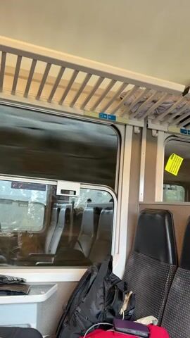 The video captures the view from inside a moving train. The interior is visible, showing rows of seats with black headrests, a luggage rack above, and a small table. A backpack and other belongings are on a seat. The train is moving quickly, as evidenced by the blurry landscape seen through the window. Outside, there are trees and mountains, partially obscured by fog or clouds. A warning sticker is visible on the window frame, advising against throwing objects out or leaning outside.