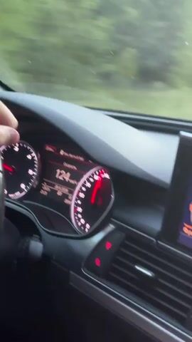 This media file captures a drive through a rural landscape in Dovhe, Ukraine. The perspective is from inside a car, showing the dashboard and the road ahead.

The dashboard displays a digital speedometer indicating a speed of 124 km/h. The car's infotainment screen is visible, showing a menu with Russian text, including options like "Копировать" (Copy) and a list of items that appear to be song titles or playlist entries such as "Мое, Хочу" (Mine, I Want) and "Молитва за" (Prayer for).

The scene transitions to the view through the windshield. It's dusk, with the sun setting and casting a warm orange glow on the horizon. The road is paved and appears to be a two-lane highway, bordered by dense trees on either side. The sky is a gradient of pale blue to orange, suggesting the end of the day. The overall mood is peaceful and serene, with the motion of the car creating a sense of journey. The weather appears clear, with no visible precipitation.