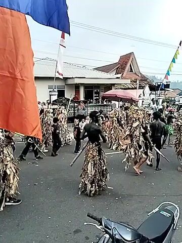 The video shows an outdoor street parade with a group of people in elaborate costumes. Many individuals are dressed in full-body coverings made of dried leaves or similar plant material, giving them a unique, earthy appearance. These costumed figures appear to be marching in formation on a paved road. In the background, buildings, flags (including an Indonesian flag), and spectators can be seen. There are also some individuals not in elaborate costumes, including what appear to be children in bright, fringed outfits participating in the parade. Some of the leaf-costumed individuals seem to be carrying staffs or similar props. The overall atmosphere suggests a cultural or festive event.