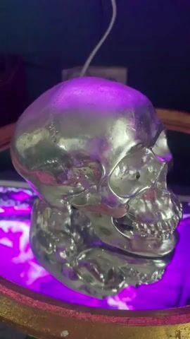 The video shows a close-up, side view of a shiny, metallic skull. The skull is resting on a reflective surface, possibly a mirror, which is illuminated by a purple light. The background is dark and out of focus, with hints of other objects and a dark blue wall. A thin white wire is visible hanging from above the skull. The lighting creates a dramatic and somewhat eerie atmosphere.