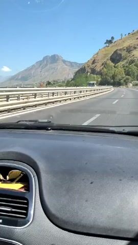 The video shows a first-person view from inside a car driving on a highway. The road has multiple lanes and is bordered by a guardrail. On the left side of the road, there is a large mountain in the distance with a clear blue sky above. On the right side, there is a hillside covered in greenery. Other vehicles, including a bus, are visible on the highway. The car's dashboard is in the foreground, with a pair of sunglasses resting in a compartment. The overall impression is of a scenic drive through a mountainous region on a sunny day.