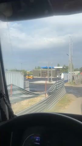 A yellow steamroller is seen compacting asphalt on a construction site. The driver is visible operating the machine. In the background, there's a building with scaffolding and construction materials. The sky is cloudy. The perspective is from inside a vehicle, looking out through the windshield.