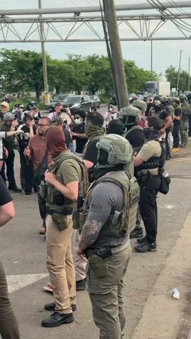 The video shows a group of people standing on a sidewalk. Most of them appear to be law enforcement officers or security personnel, as they are wearing tactical gear, helmets, and some have visible firearms. Some of them have masks or face coverings. There are also several people who look like onlookers or reporters, taking pictures with their phones or cameras. The background includes cars, trees, and utility poles. In the background, someone can be heard shouting. There is trash on the side of the road.